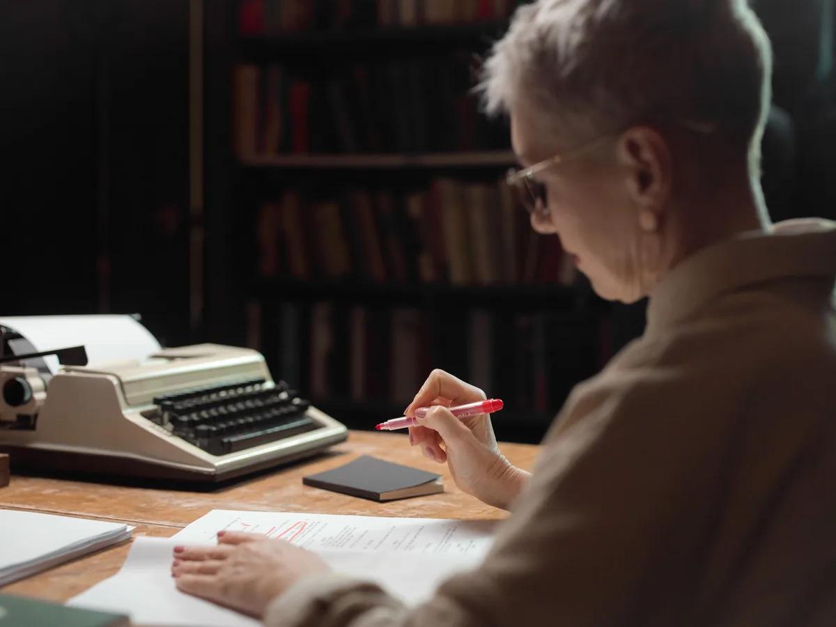 Someone marking up a typed page in red pen beside a typewriter, in front of bookshelves
