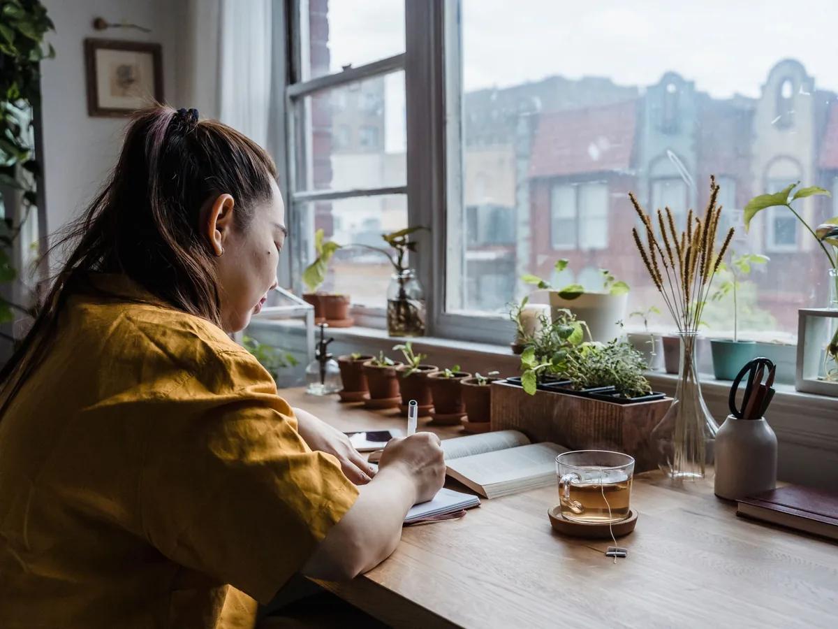 Someone writing in a notebook at a plant-filled desk in front of a window