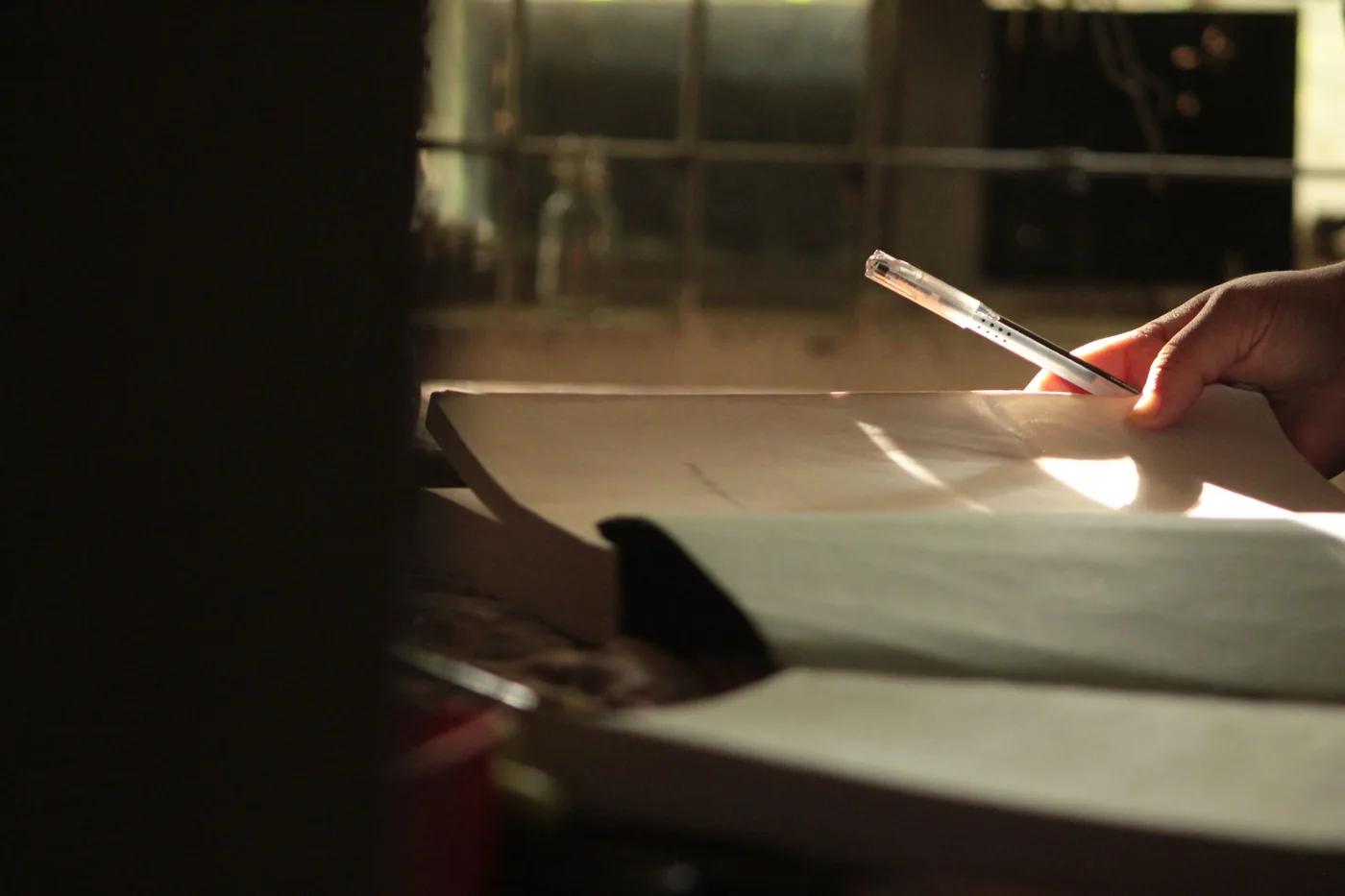 A hand writing on paper at a table in warm light from a nearby window