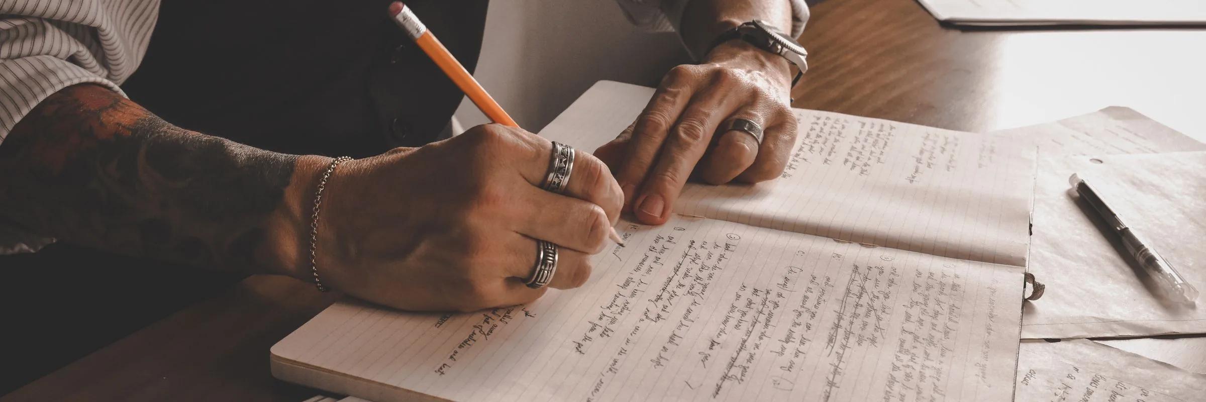 Close view of two hands writing in pencil across a notebook filled with handwritten pages